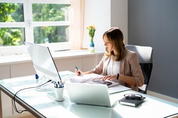 Mulher com uma calculadora, notebook e documentos em cima de uma mesa anotando algo com uma caneta