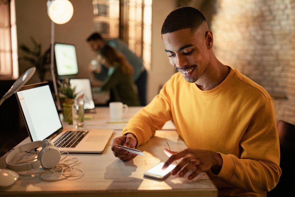 Homem mexendo em celular com cartão na mão e notebook